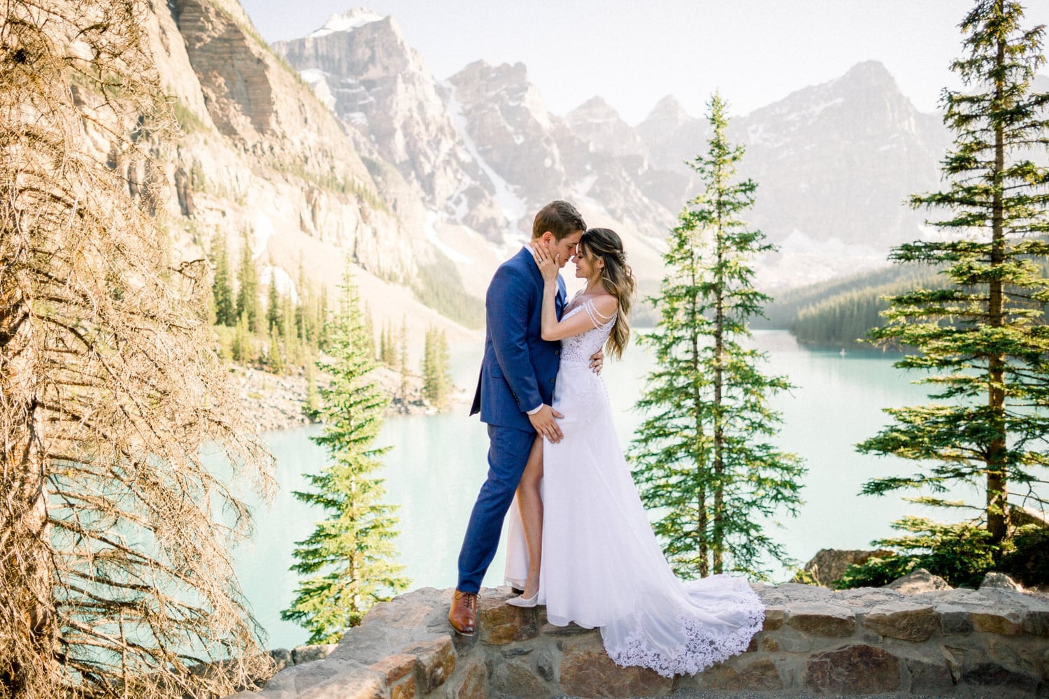 A bride and groom share a tender kiss by a serene lake surrounded by mountains, capturing a moment of love in nature.