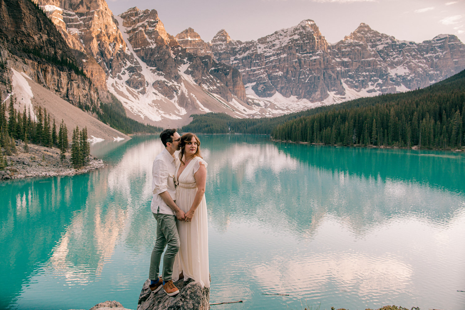 A couple stands close together on a rock by a serene turquoise lake, surrounded by majestic mountains and lush forests, capturing a moment of intimacy and love.