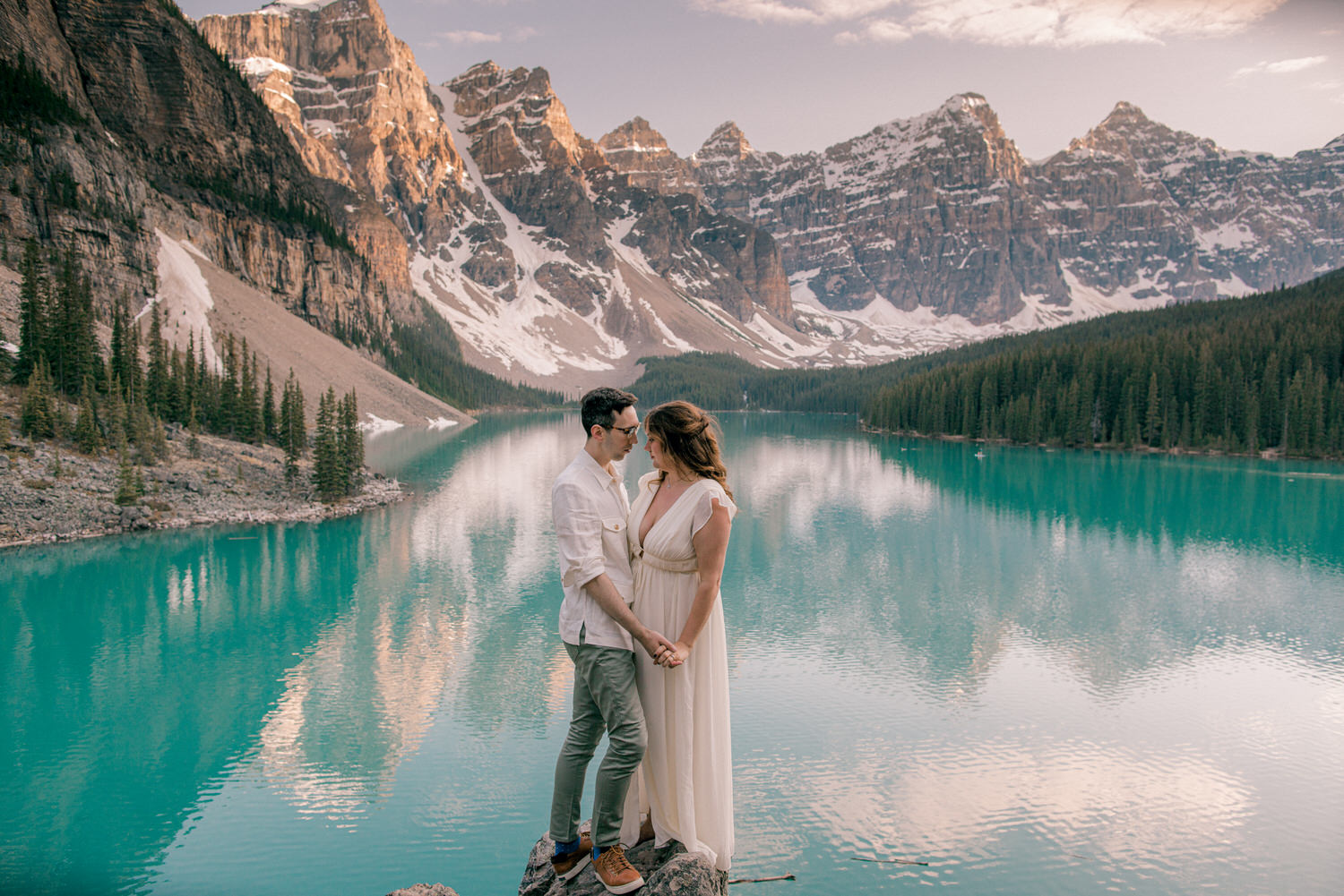 A romantic scene with a couple standing on a rock, holding hands by a serene turquoise lake surrounded by majestic mountains and evergreen trees, during golden hour.