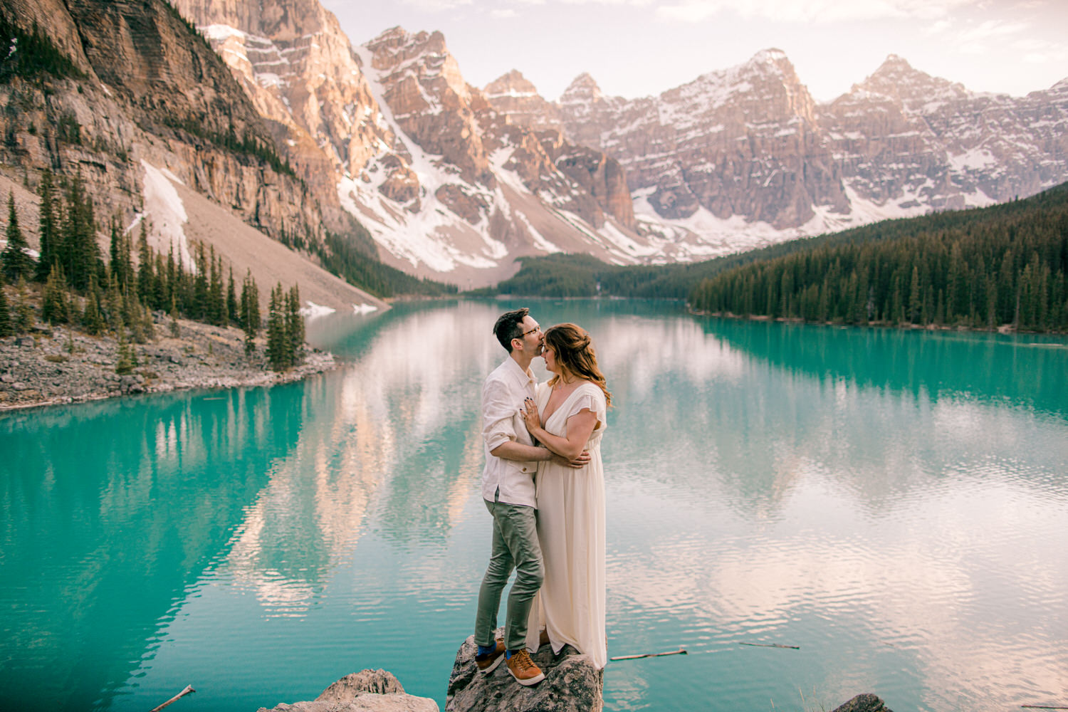 A couple sharing a tender moment by a stunning turquoise lake, surrounded by majestic mountains and a serene forest backdrop.