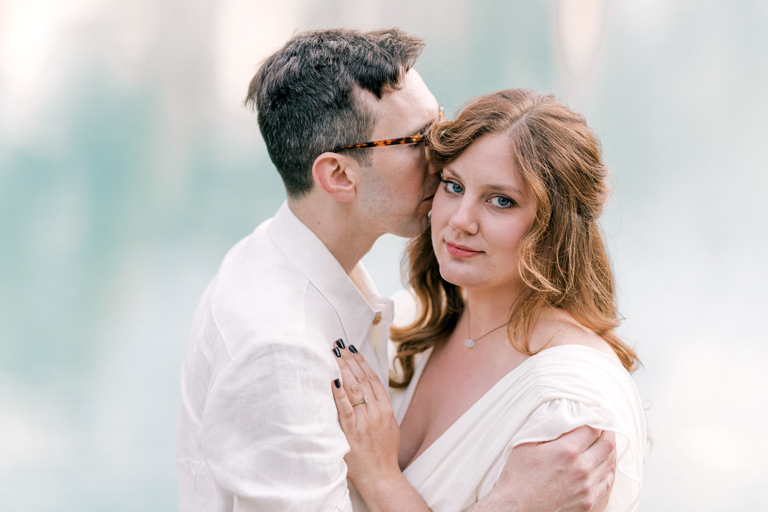 A couple shares a tender moment by a serene water backdrop, with the man gently kissing the woman's cheek as she gazes softly at the camera.