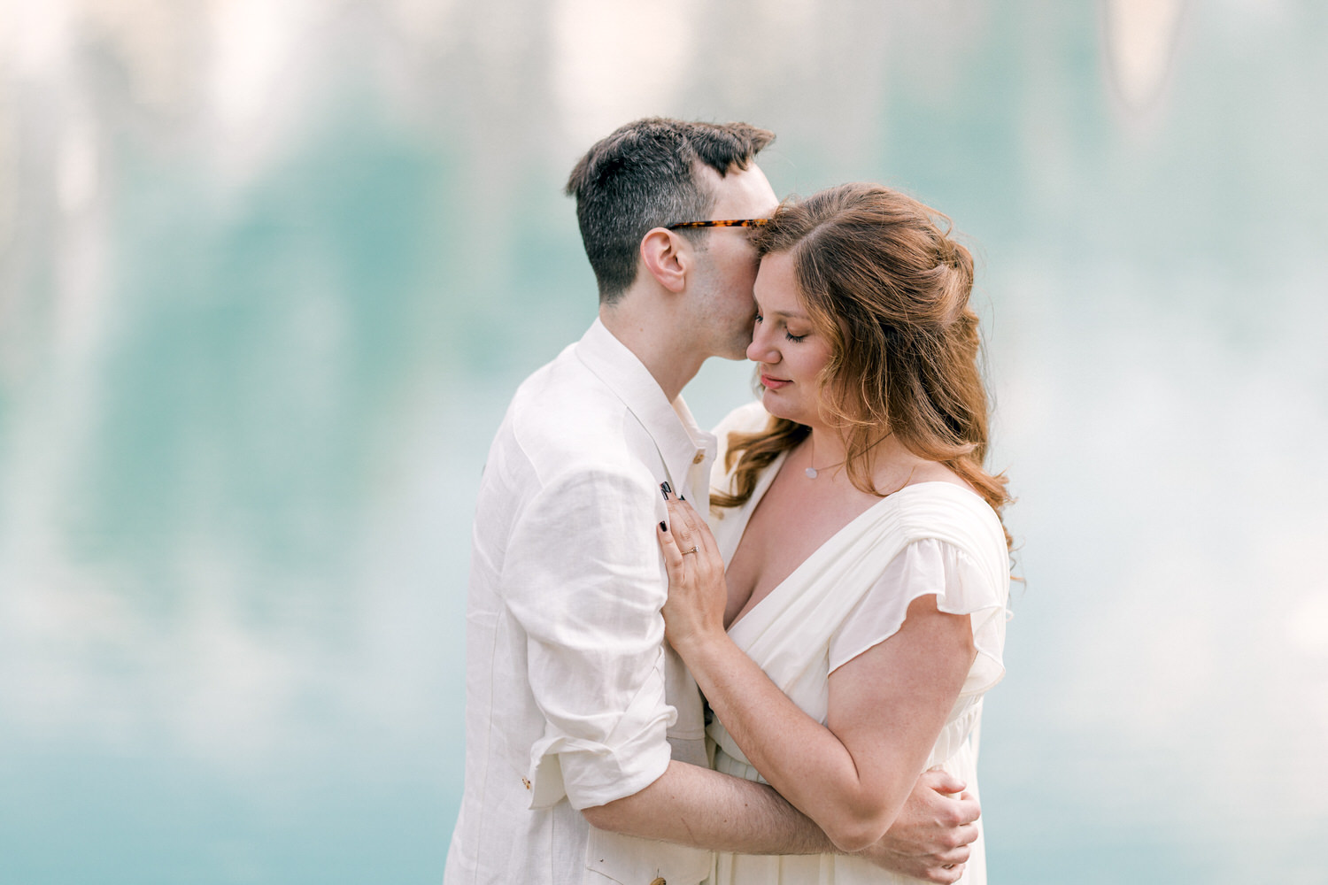 A loving couple sharing an intimate moment by a serene water backdrop, with the man gently kissing the woman's forehead.