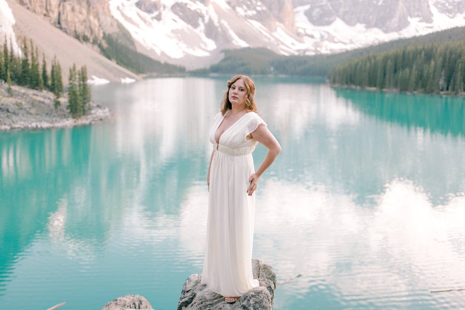 A woman in a flowing white dress stands confidently on a rock by a tranquil turquoise lake, surrounded by towering mountains and lush evergreen trees.