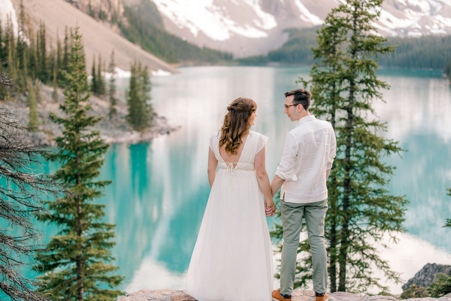 A couple holding hands, gazing at a scenic turquoise lake surrounded by mountains and trees. The woman wears a flowing white dress, while the man is dressed in a light shirt and green pants.