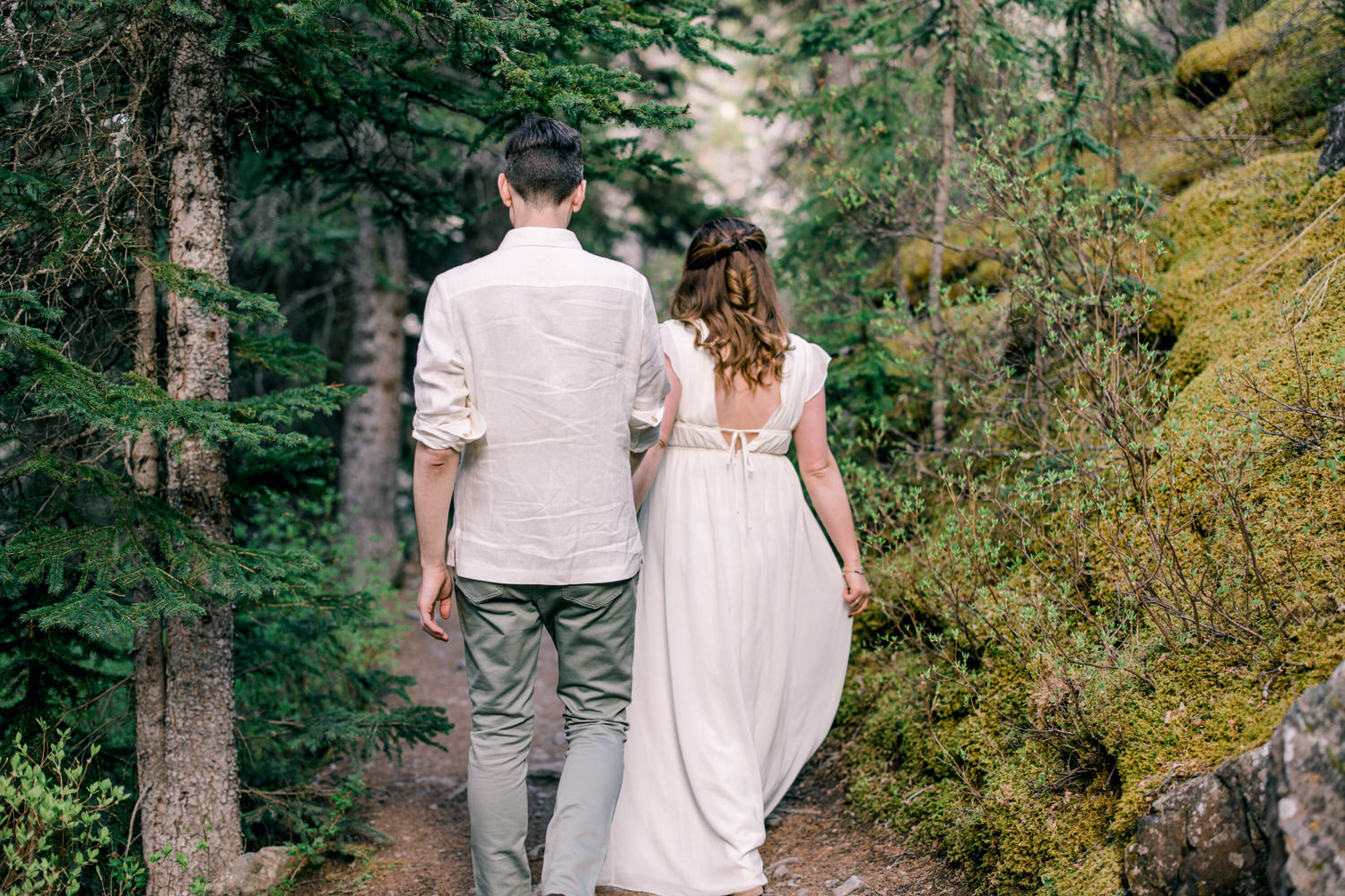 A couple walking hand in hand along a wooded trail, surrounded by lush greenery and tall trees, exuding a romantic and tranquil atmosphere.