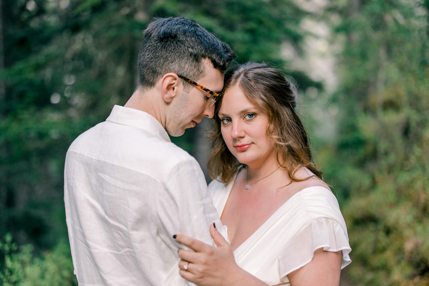 A couple embraces in a serene forest setting, with the woman gazing directly at the camera while the man rests his forehead against hers.