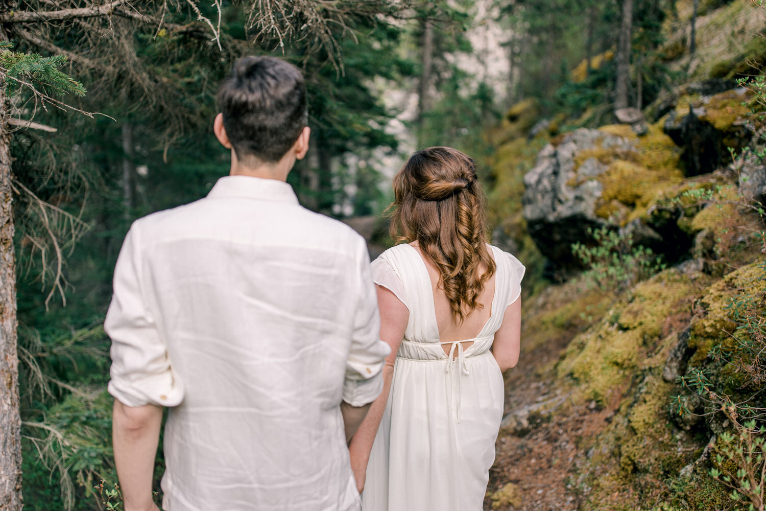 A romantic couple in a forest, the woman in a flowing white dress and the man in a light shirt, walking hand in hand along a moss-covered path surrounded by trees.