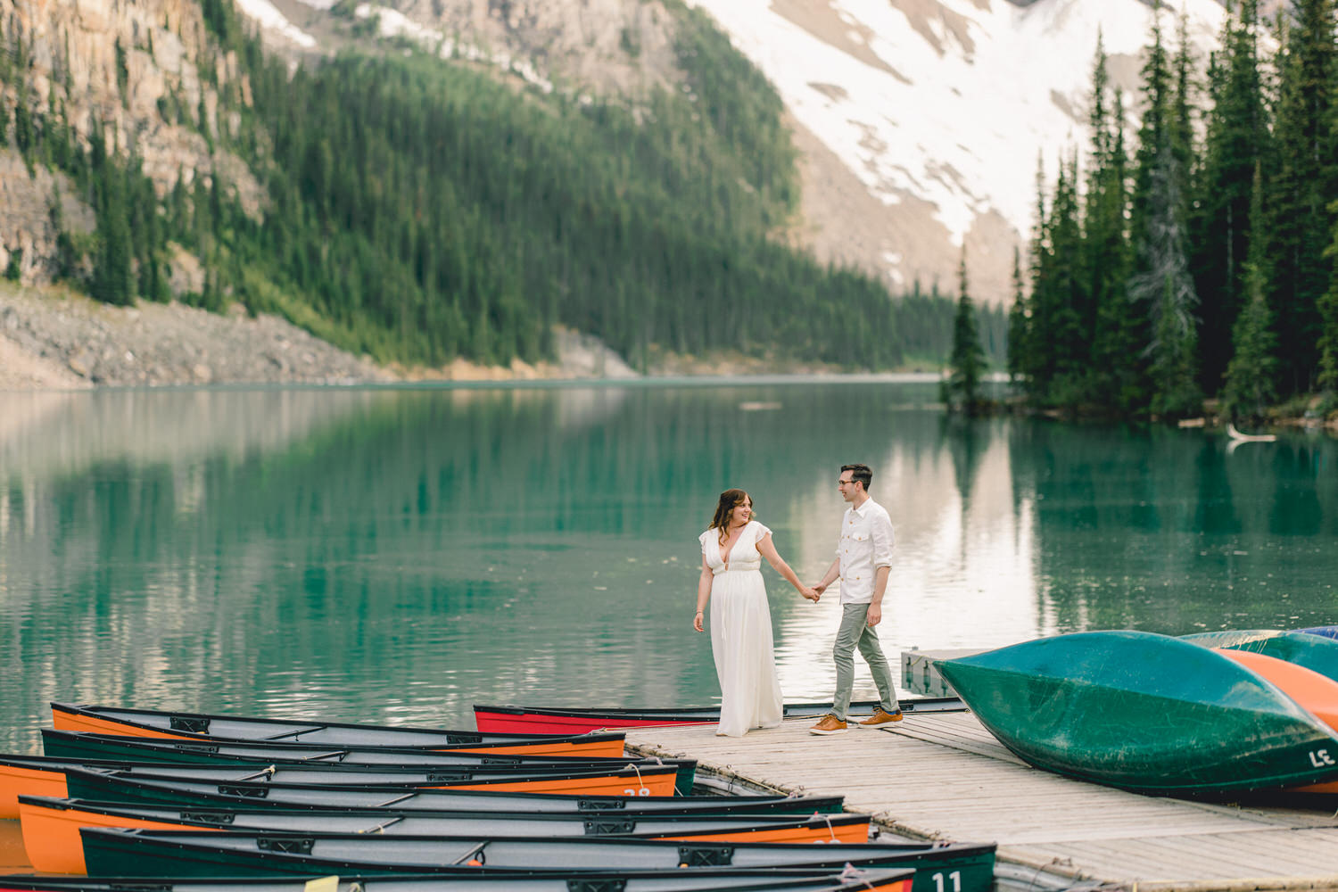 Couple Embracing Nature::A couple holding hands on a dock by a serene lake, surrounded by canoes and lush green mountains.