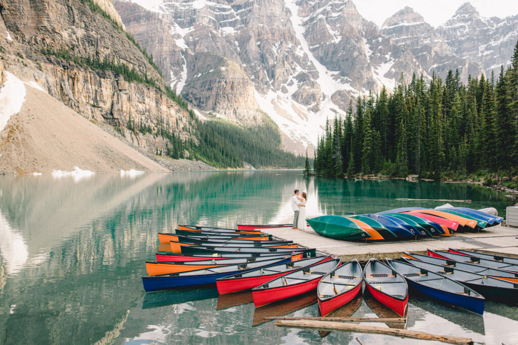 A couple embraces on the shore of a serene lake, surrounded by colorful canoes and stunning mountain scenery.