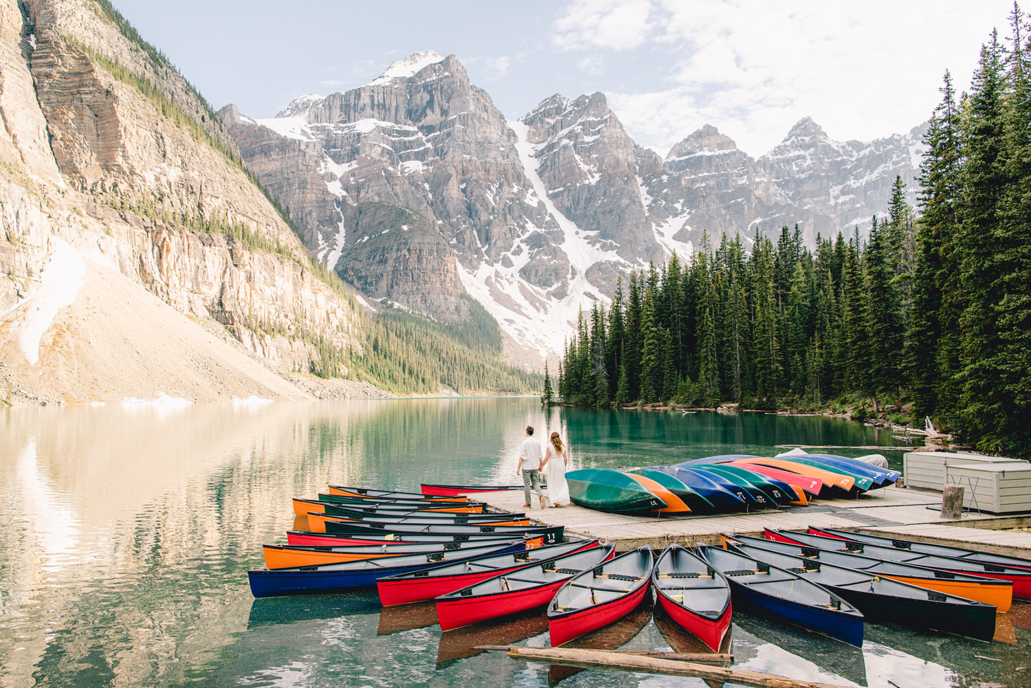 A couple walks hand in hand by a tranquil lake surrounded by colorful canoes and majestic mountains.