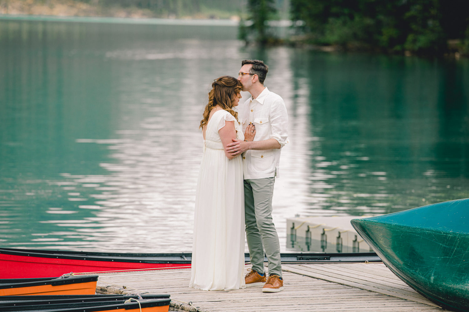 Couple sharing an intimate kiss on a wooden dock by a calm lake, surrounded by colorful boats and lush greenery.