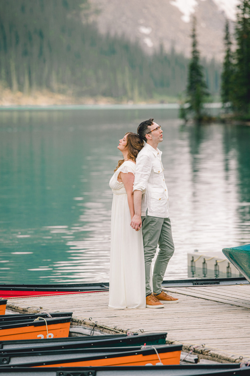 A couple standing back-to-back on a dock by a tranquil lake, holding hands, surrounded by lush trees and reflecting waters.