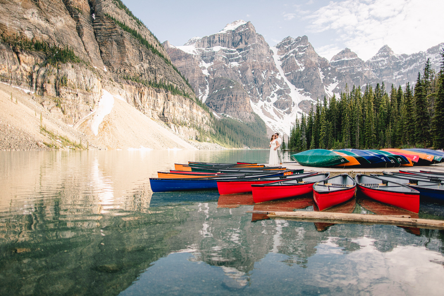 A couple embraces on the shore near a cluster of colorful canoes, surrounded by majestic mountains and pristine waters.