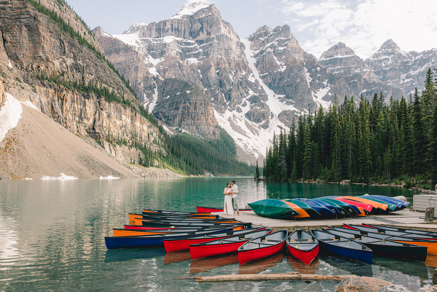 A couple embraces on a dock surrounded by colorful canoes, with majestic mountains and lush forests reflecting in the serene water.