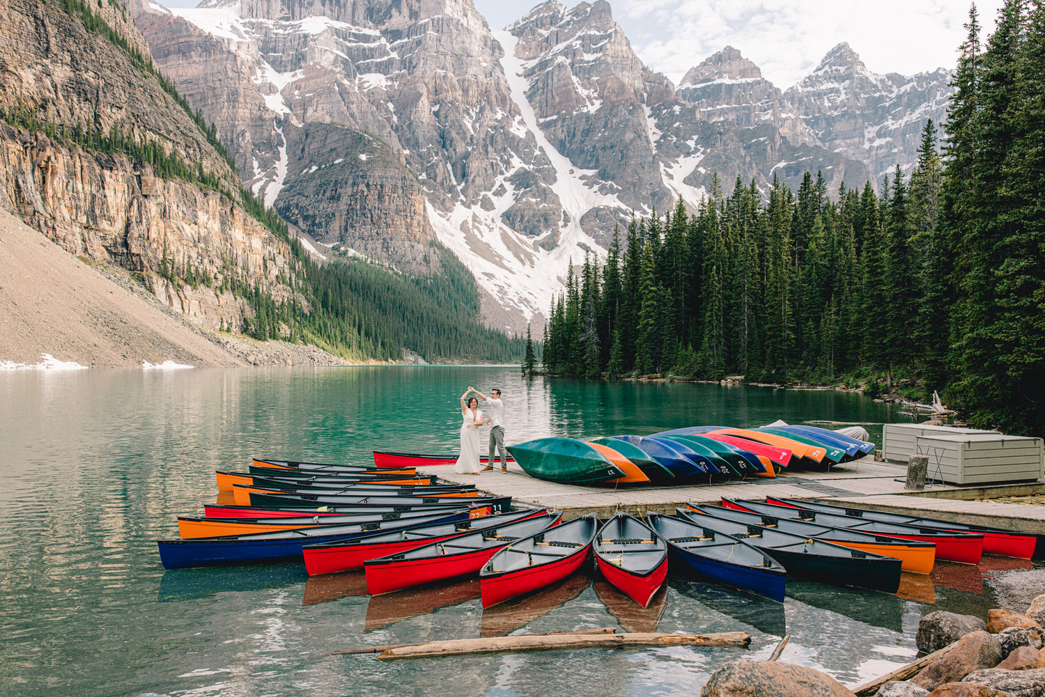 A couple dances on a dock surrounded by colorful canoes, with majestic mountains and a serene lake in the background.