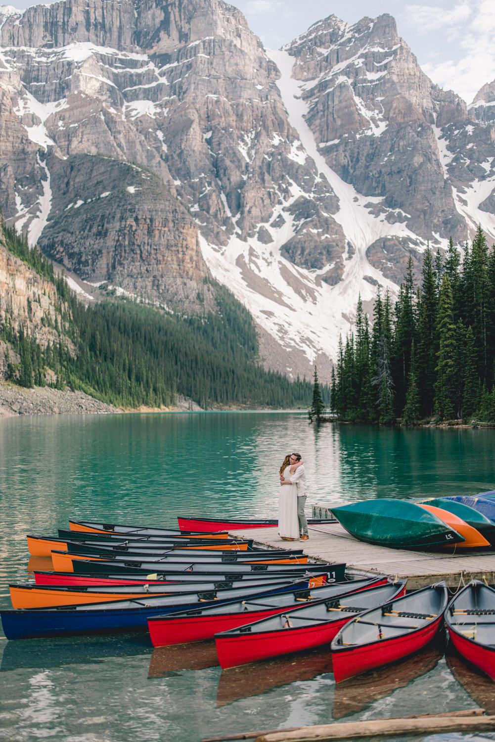 A couple shares a tender moment on a dock surrounded by colorful canoes and stunning mountain scenery with snow-capped peaks.