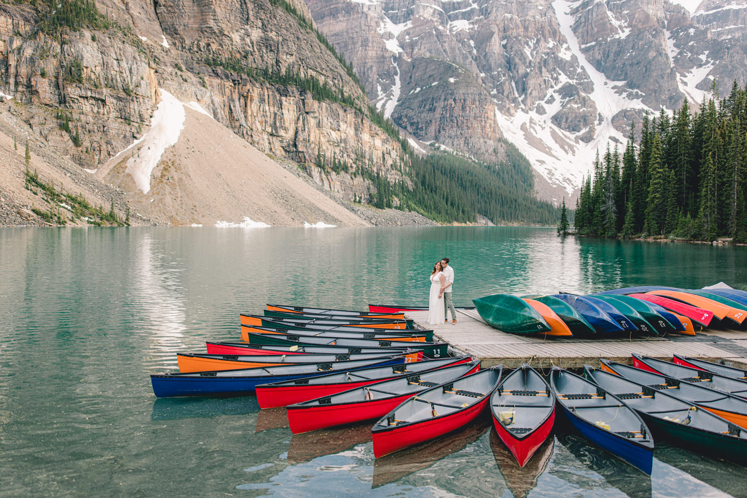 A couple embraces on a wooden dock surrounded by colorful canoes with serene mountains and a turquoise lake in the background.