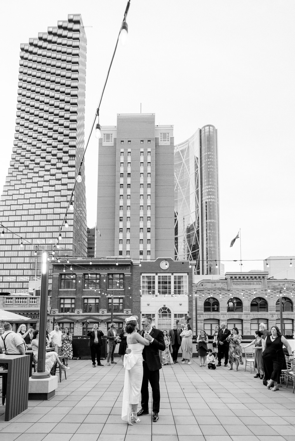 Couple dancing elegantly on a rooftop with city skyline and guests watching, adorned with string lights.