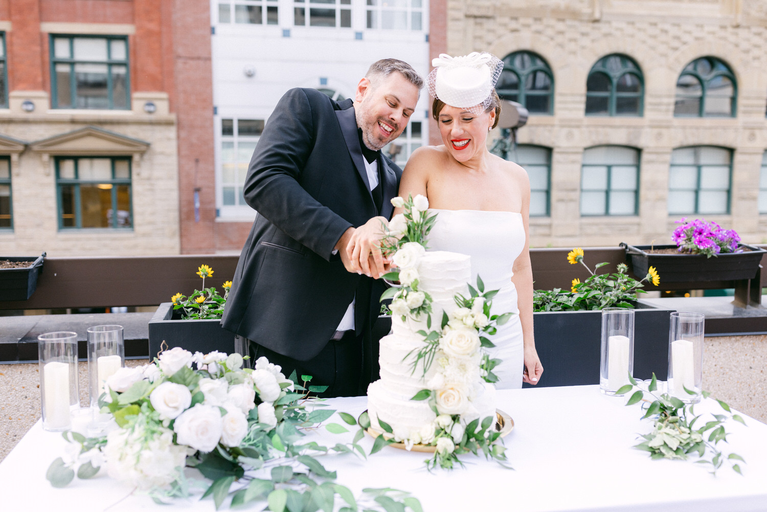 A couple joyfully cutting a beautifully adorned wedding cake on a rooftop, surrounded by floral decorations and elegant candles.