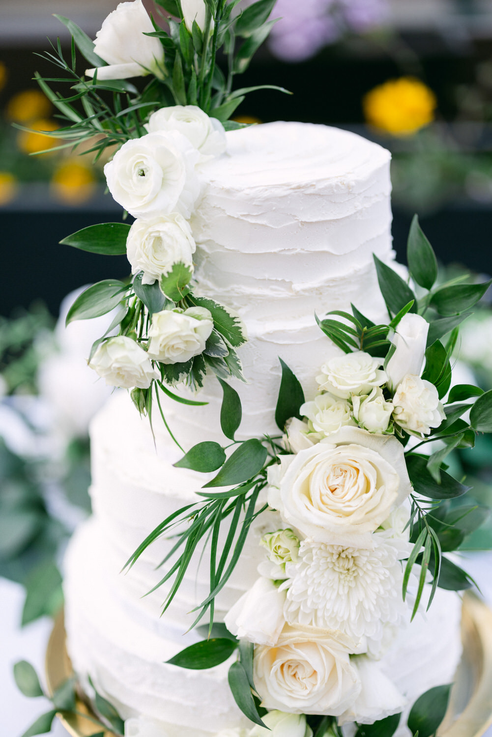 A three-tiered white wedding cake adorned with fresh white roses and greenery, set against a blurred floral background.