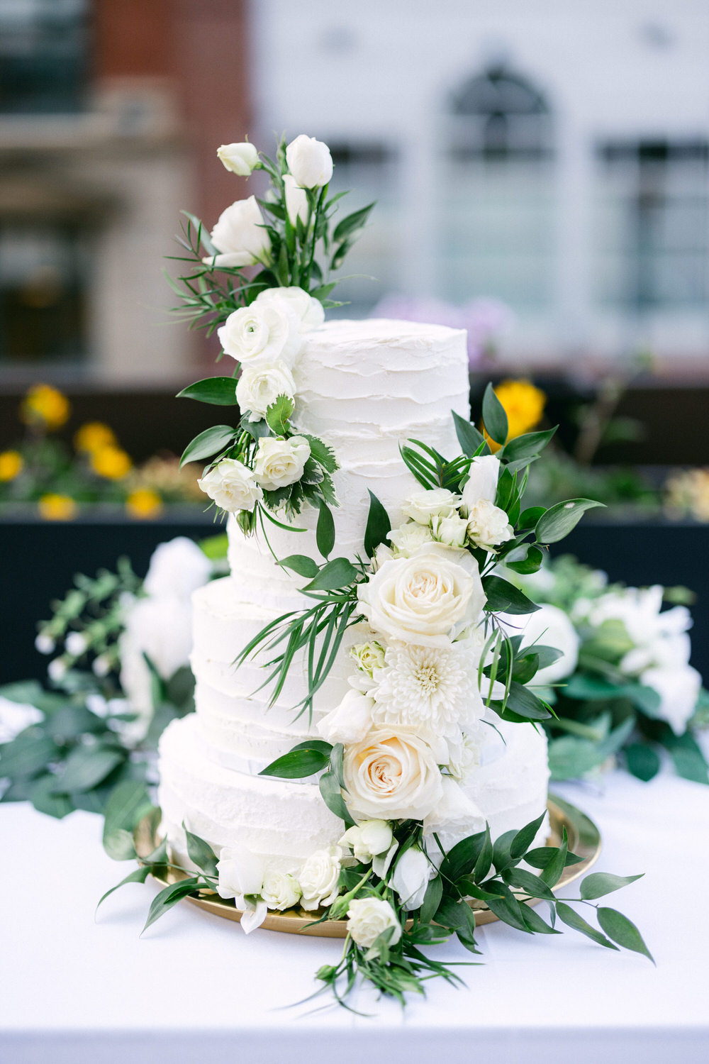 A three-tiered wedding cake adorned with white roses and greenery, set on a golden platter against a soft-focus garden backdrop.