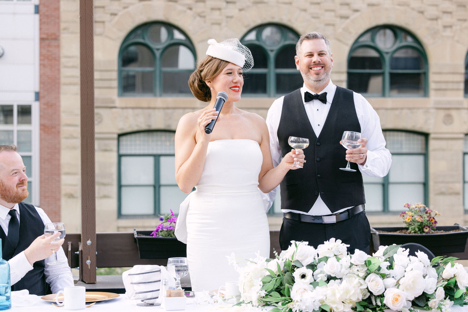 A bride delivering a toast with a microphone, accompanied by a groom holding a champagne glass, all set against a backdrop of elegant floral arrangements and a charming cityscape.