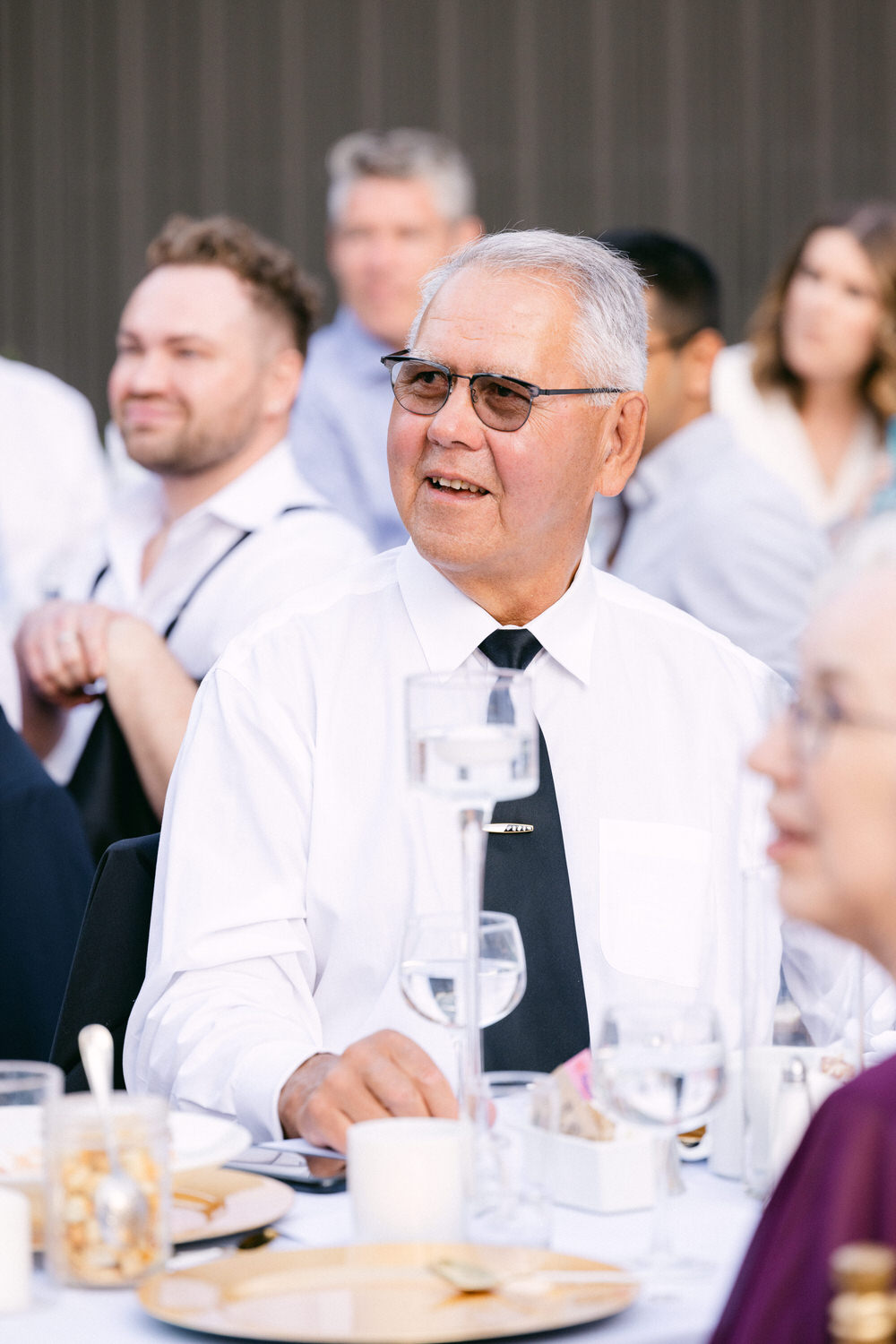 An older man in a white dress shirt and black tie smiles while sitting at a table with guests during a celebration, surrounded by glasses of water and plates.