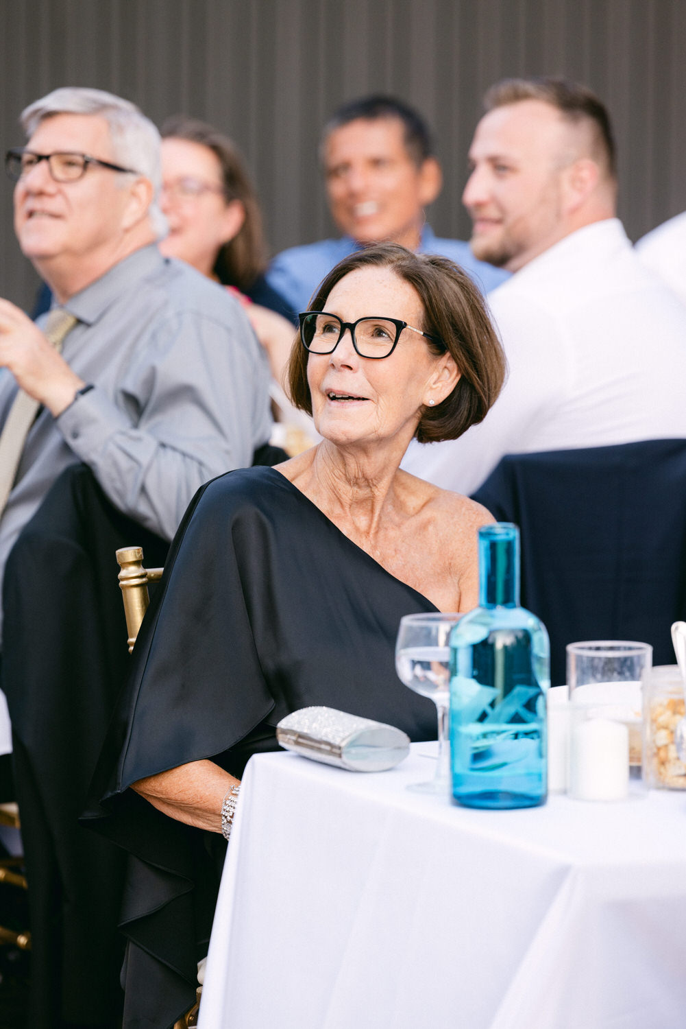 A woman in a black dress enjoys a lively event, seated at a table with a blue bottle and glasses, surrounded by smiling guests in the background.