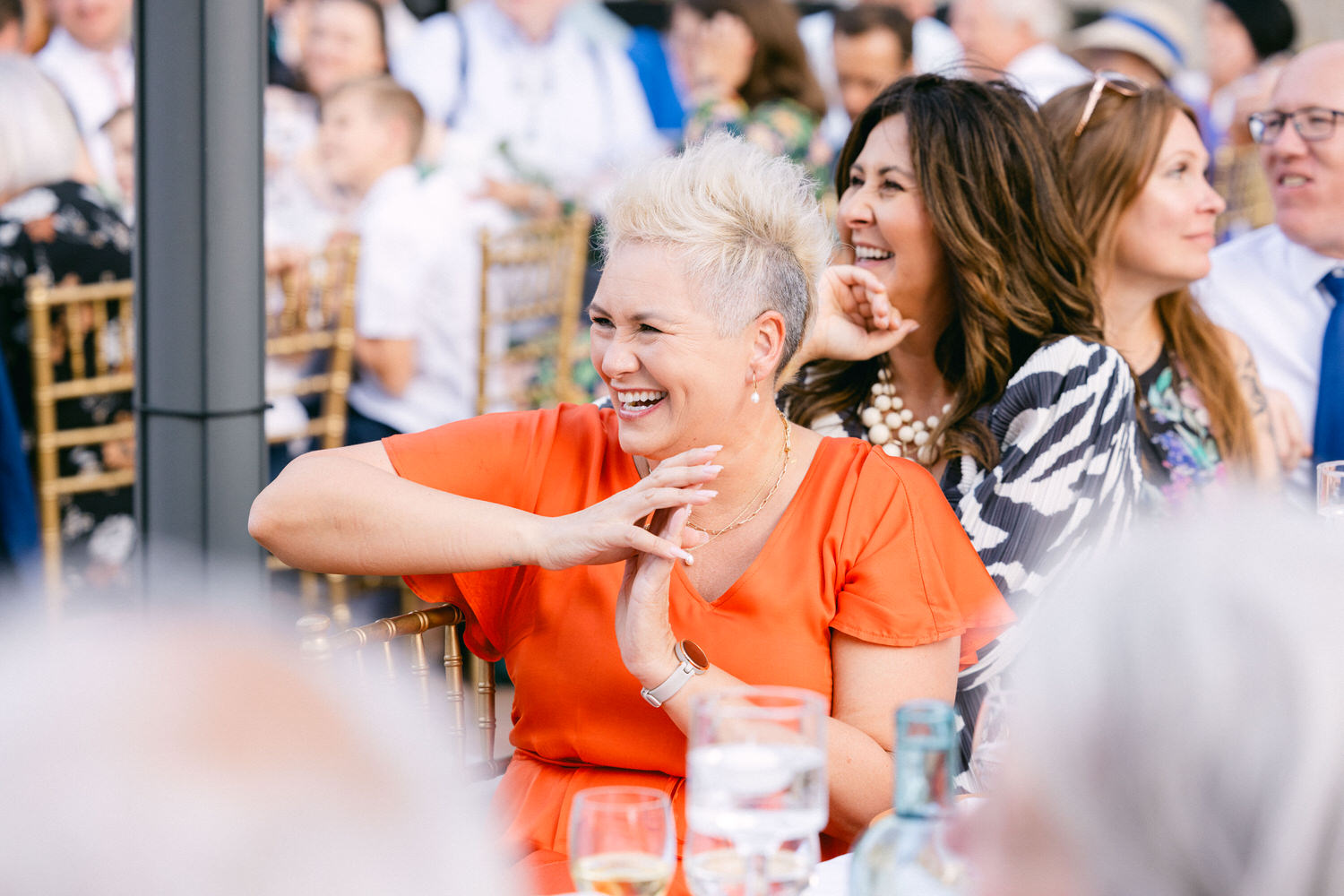 A woman in a vibrant orange dress laughs joyfully at a gathering, surrounded by friends and family, with glasses of drinks on the table.