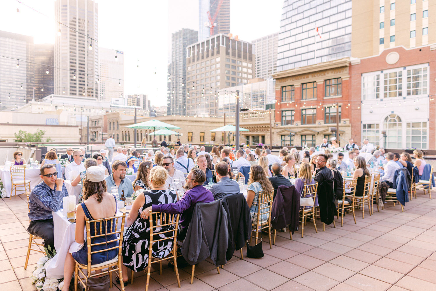 A lively outdoor dining setup with guests seated around a long table, enjoying a gathering against a backdrop of city skyscrapers, with string lights overhead.