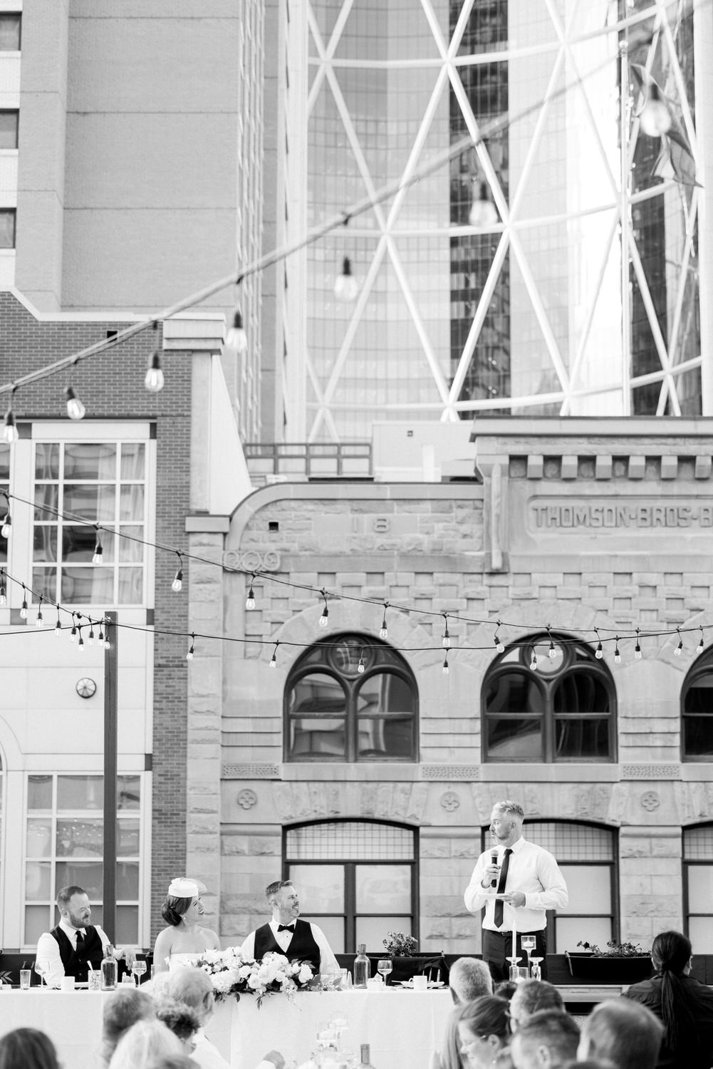 A groom delivering a heartfelt toast at a wedding reception, with guests seated at a long table adorned with floral decor, against a backdrop of modern and historic buildings.