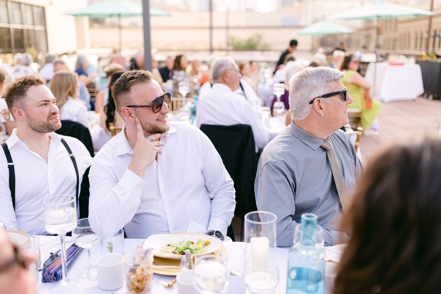 A group of men seated at a dining table outdoors, with one man in sunglasses looking thoughtfully, enjoying their meal against a backdrop of a bustling event.