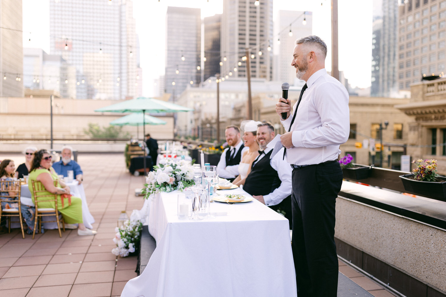 A speaker addresses wedding guests seated at a beautifully decorated outdoor table with city skyscrapers in the background.