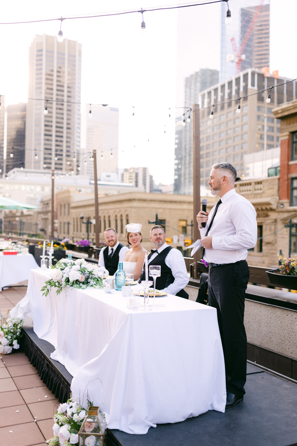 A man speaking at a wedding reception with the bride and groom sitting at a beautifully decorated table surrounded by urban scenery.