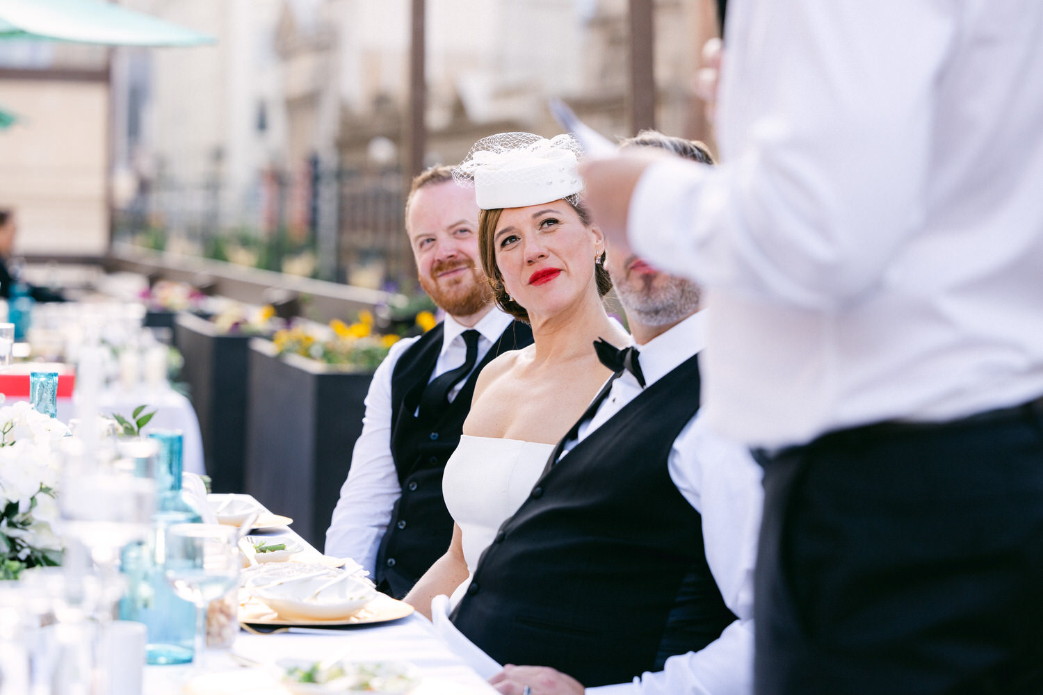 A joyous outdoor wedding reception featuring a bride in a stylish white dress and feathered hat, surrounded by guests in formal attire at a beautifully set table.