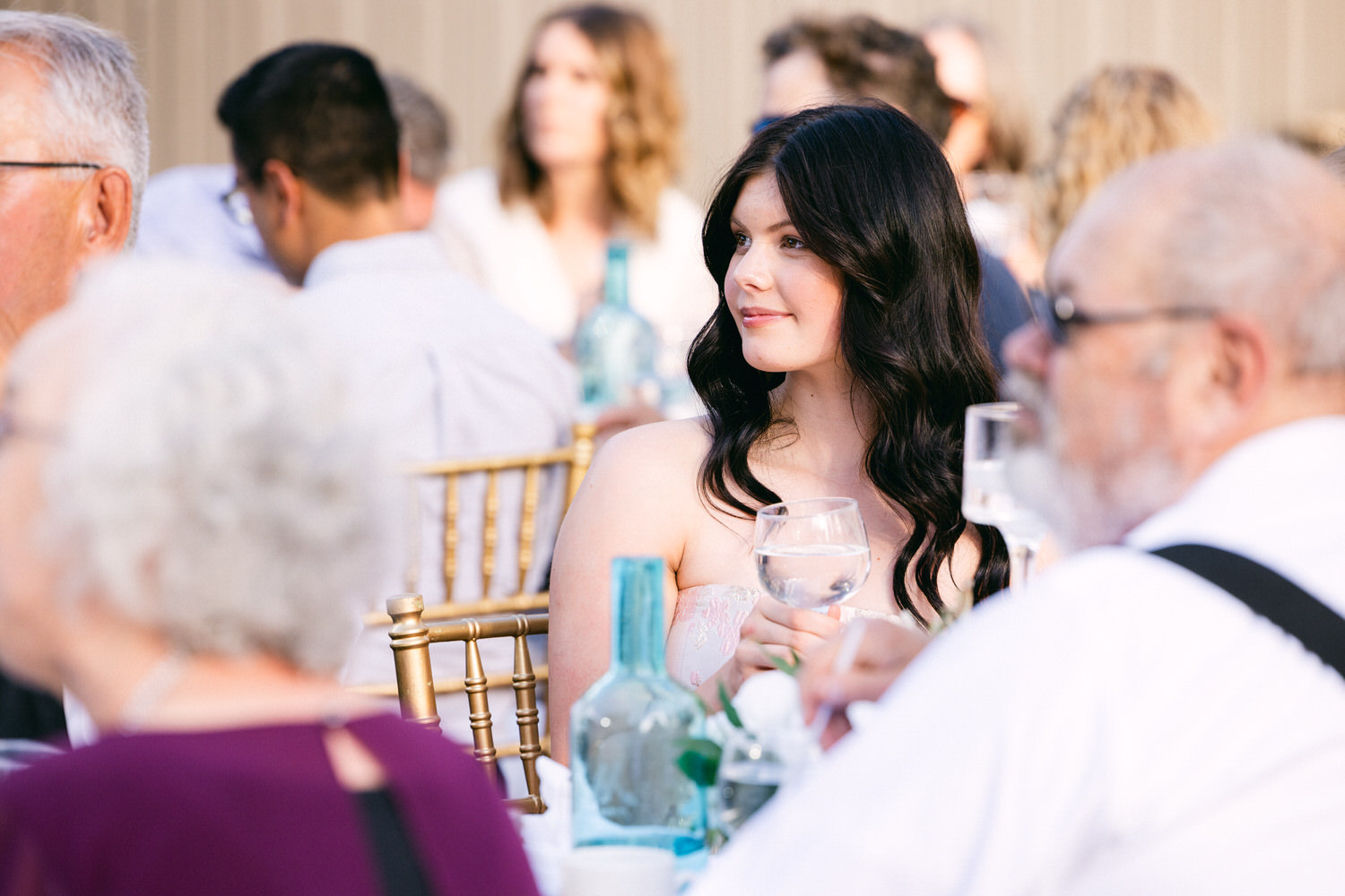 A young woman in a beautiful dress holds a glass of water while sitting among guests at a stylish outdoor event.