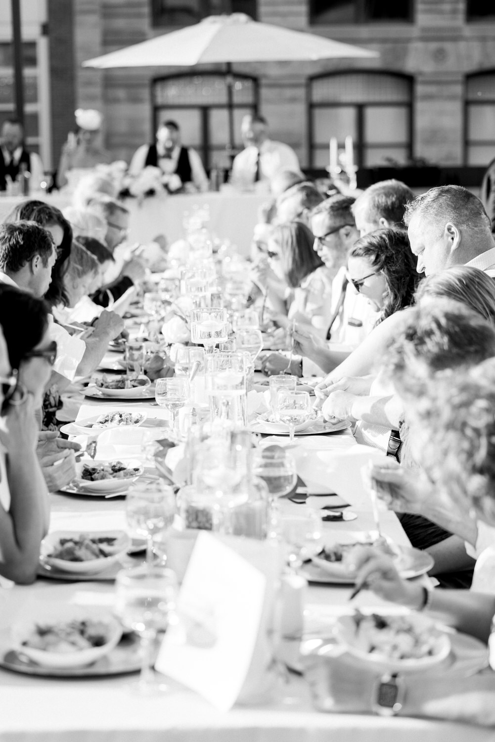 A long table filled with guests enjoying a meal outdoors, surrounded by drinks and plates of food, in a sunlit setting.