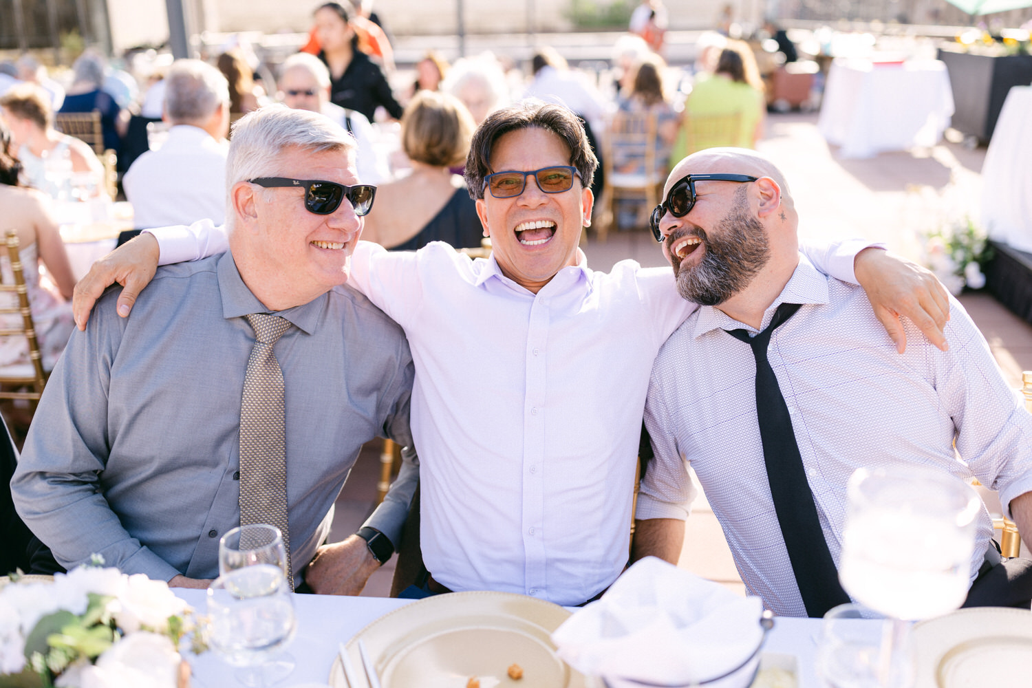 Three men laughing and enjoying a sunny outdoor celebration, sitting close together at a decorated table with drinks, surrounded by a cheerful crowd.