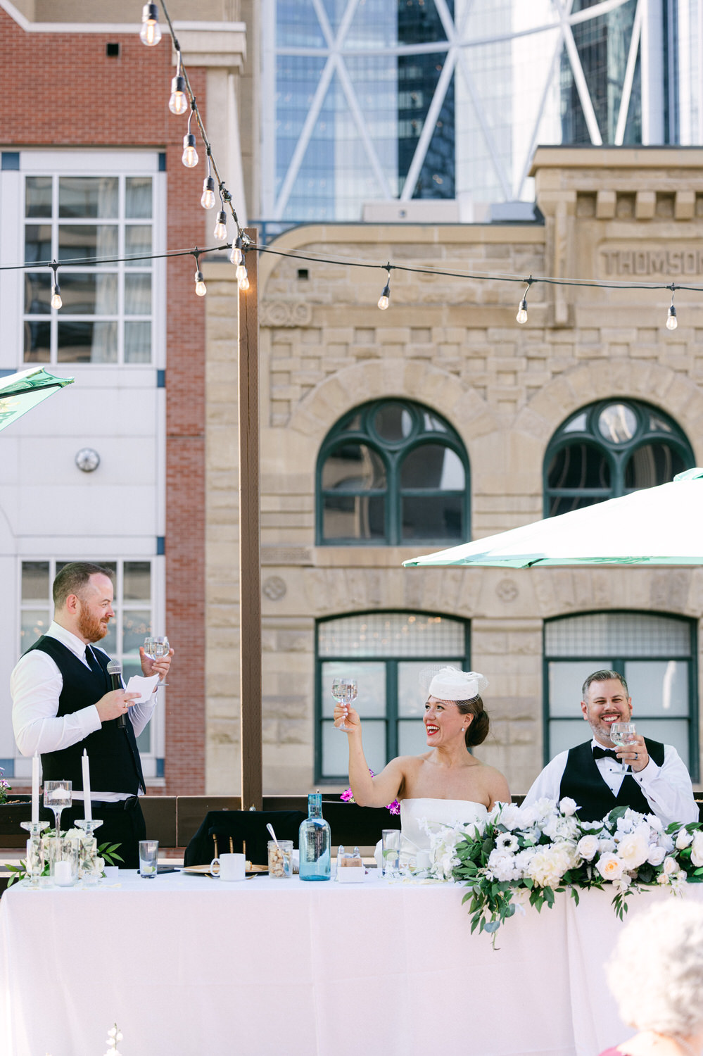 A bride and groom toast during their wedding reception, surrounded by elegant decor and a city backdrop.