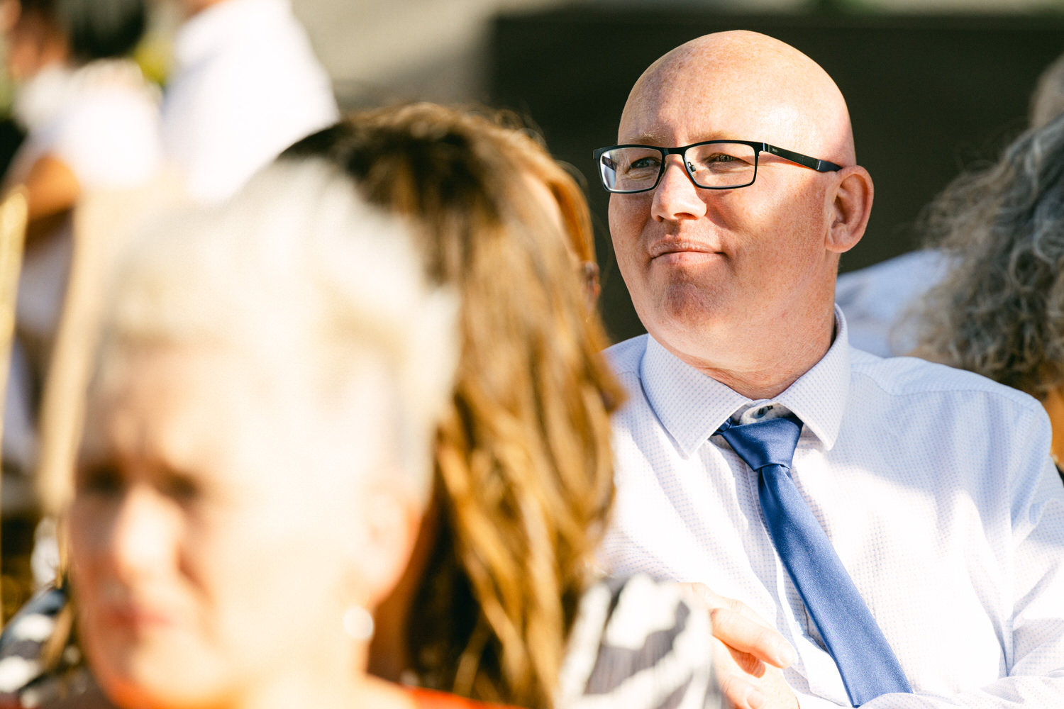 A bald man wearing glasses and a blue tie, sitting among other attendees, appears engaged during a gathering.