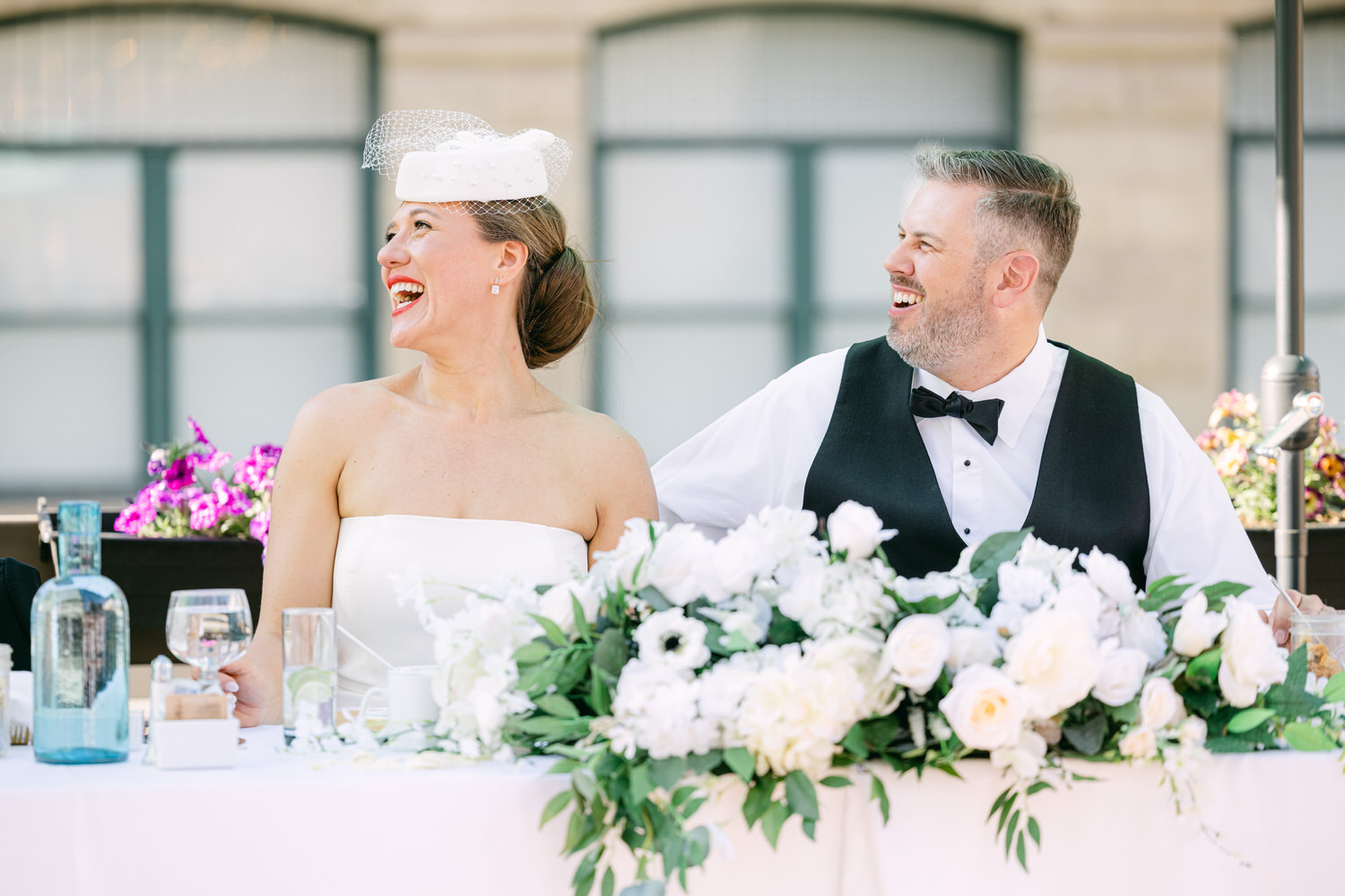 A smiling bride and groom share a lighthearted moment during their wedding reception, surrounded by elegant floral decorations and drinks.