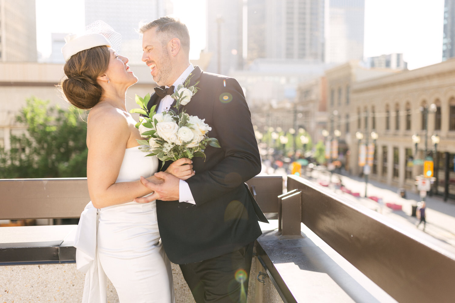 A joyful couple shares a moment on a balcony, with the city skyline in the background and the bride holding a bouquet of white roses.