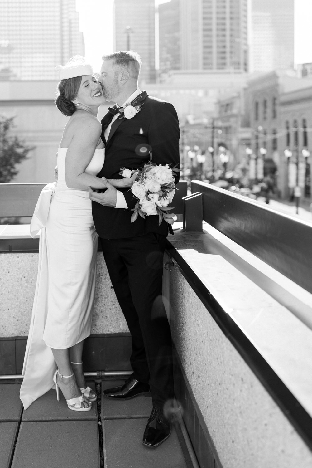 A joyful couple shares an intimate moment on a rooftop with city buildings in the background, showcasing a beautiful bouquet and formal attire.