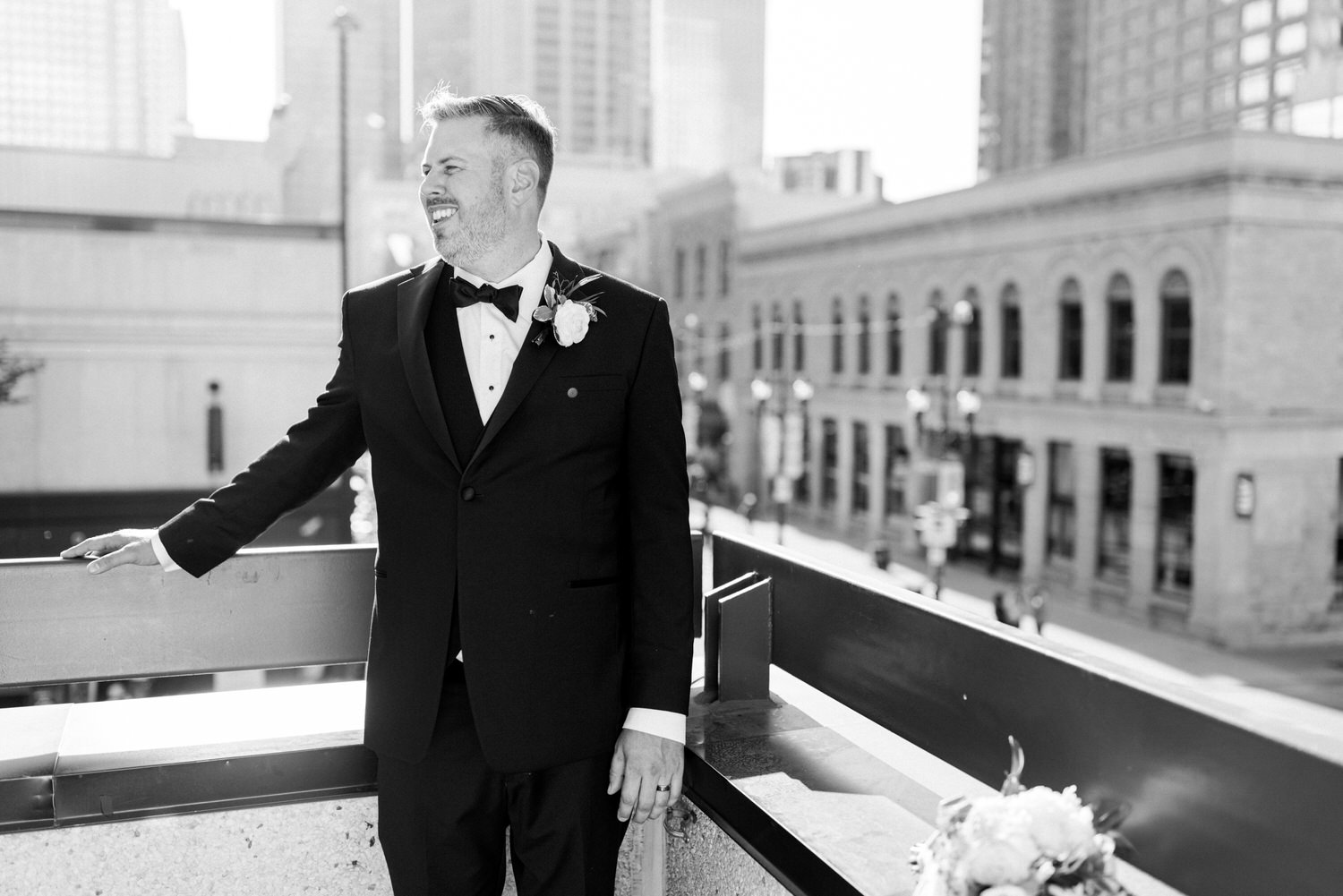 A smiling groom in a black tuxedo stands on a balcony with a cityscape in the background, holding onto the railing.