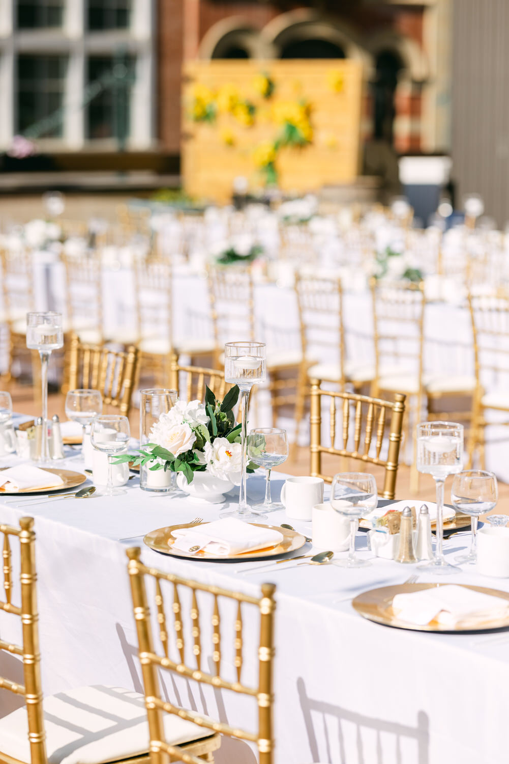 A beautifully arranged outdoor dining area featuring gold chairs, white tablecloths, floral centerpieces, and elegant dining ware, set for an event in a sunny atmosphere.