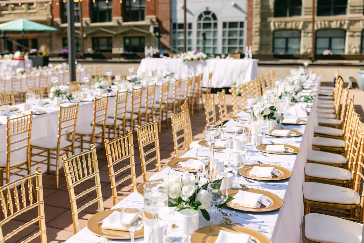 A beautifully arranged outdoor wedding reception featuring long tables with gold chairs, white tablecloths, elegant floral centerpieces, and glassware, set against a picturesque backdrop.