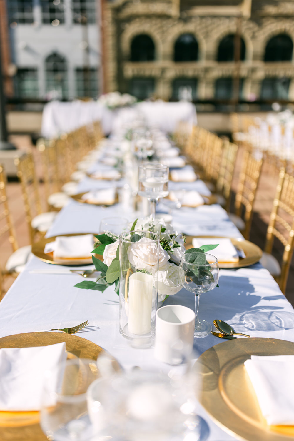 A beautifully arranged long dining table adorned with white and gold tableware, elegant floral centerpieces, and surrounded by golden chairs, set in a sunny outdoor environment.