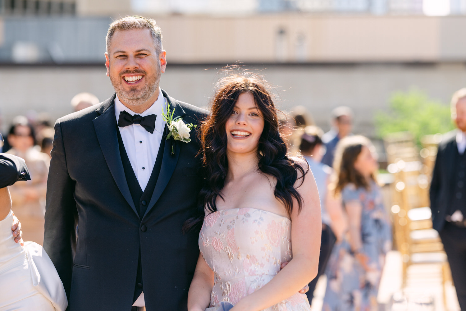 A joyful couple smiles together during a wedding ceremony, surrounded by guests in a sunny outdoor setting.