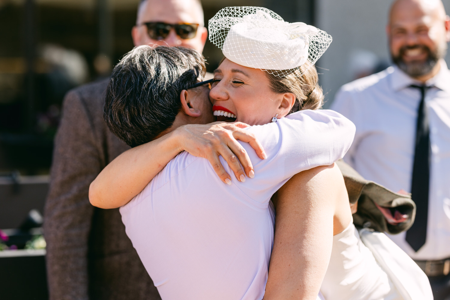 A woman in a wedding dress embraces a man, both smiling joyfully, with friends in the background enjoying the moment.