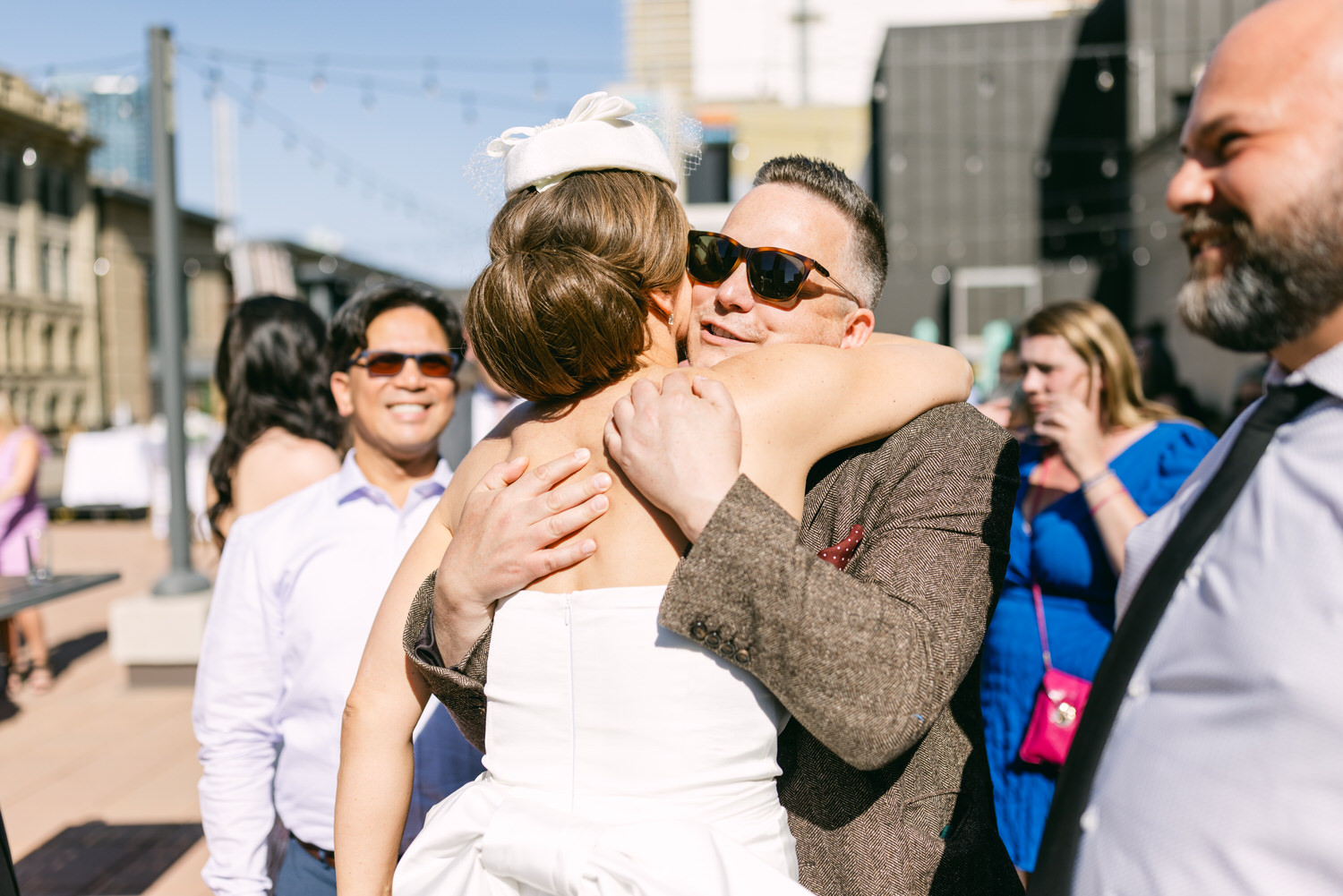 A heartfelt hug between a smiling couple, surrounded by friends at a sunny outdoor event.